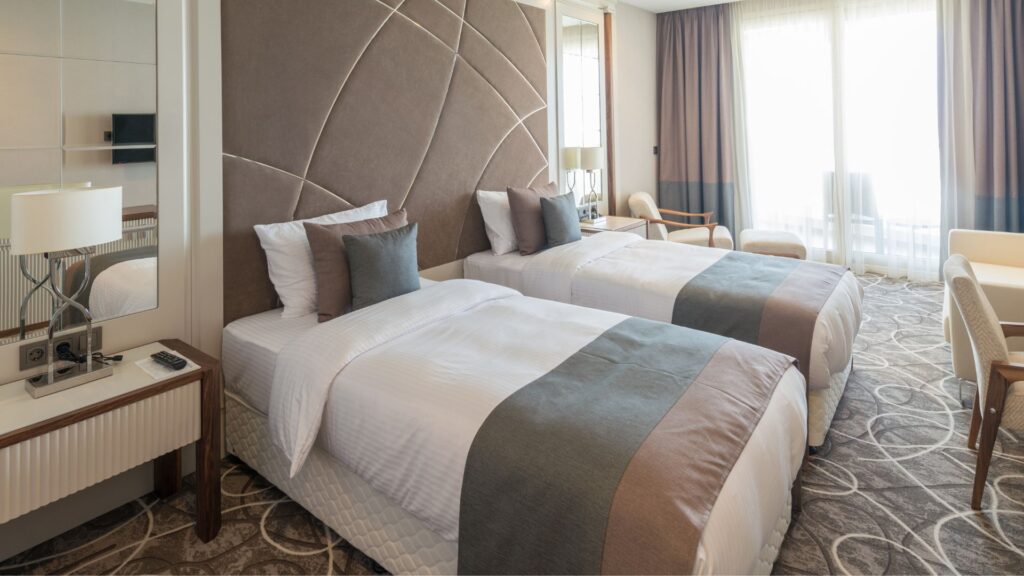 Spacious room with 2 multicolored twin beds and several matching chairs, on top of patterned carpets.